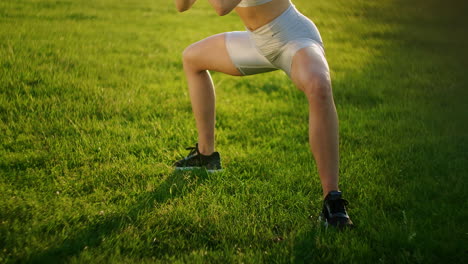 A-young-woman-performs-a-static-exercise-on-the-grass-for-the-leg-and-hip-muscles.-To-rise-on-toes-while-sitting.-Exercise-for-the-lower-leg.-Training-on-the-street-in-the-Park